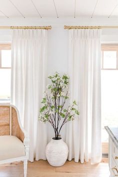 a white vase with flowers in it sitting next to a chair and window covered by curtains