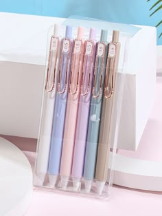 four different colored pens sitting in a clear box on a pink table next to a plant