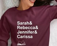 a woman laying on top of a bed wearing a maroon sweatshirt with the names of books printed on it