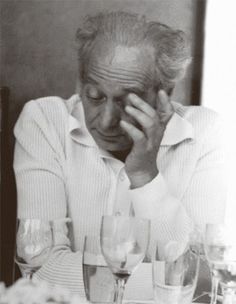 an old man sitting at a table with wine glasses in front of him and his hand on his face