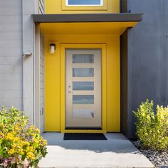 a yellow door is in front of a gray building with plants and flowers around it