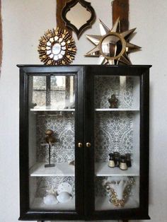 a black cabinet with glass doors and gold decorations on the top, in front of a mirror