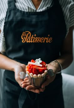 a person wearing an apron holding a cupcake with strawberries in it and marshmallows on top