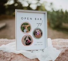 an open bar sign sitting on top of a rock next to a white sheet with three dogs