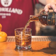 a person pouring whiskey into a glass on top of a wooden table next to oranges