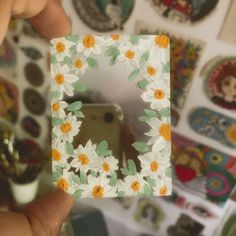 a person holding up a small square mirror with flowers on the front and sides, in front of a wall full of stickers