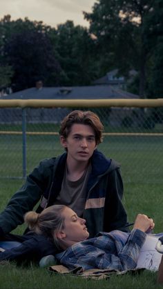 two people laying on the grass in front of a fence