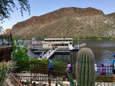 people are standing on the dock next to a river with a boat and mountains in the background