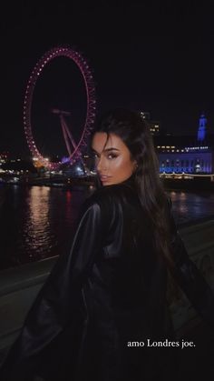 a woman standing next to the water with a ferris wheel in the background at night