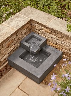 a water fountain sitting on top of a stone wall next to purple flowers and greenery