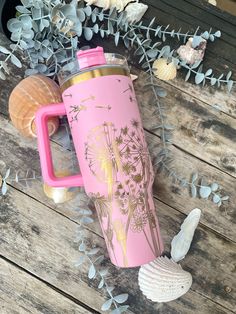 a pink and gold travel mug next to sea shells, seashells and plants