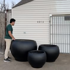 a man standing next to three large black vases in front of a white house