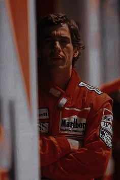 a man in an orange racing suit leaning against a pole with his arms crossed and looking off to the side