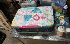 an old suitcase is covered in paint and other crafting supplies on a table next to a trash can