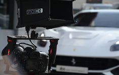 a white sports car driving down a street next to a camera on a tripod