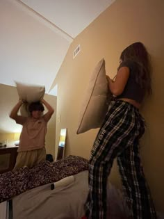 two people standing in a room with pillows on the bed and one person holding a pillow