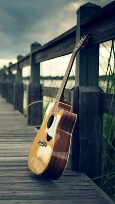 an acoustic guitar leaning against the side of a wooden fence on a pier near water