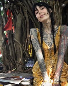 a tattooed woman sitting on the ground next to a tree with lots of books in front of her