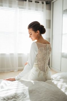 a woman sitting on top of a bed next to a window in a room with white curtains