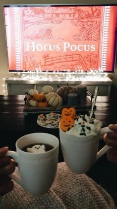 two coffee cups with marshmallows in front of a large screen television that reads hocus pocus