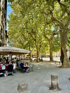 people sitting at tables under umbrellas on the sidewalk in front of trees and buildings