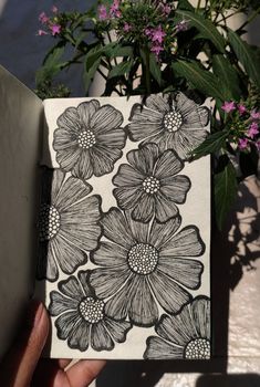 a hand holding a notebook with black and white flowers on it, next to a potted plant