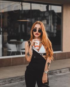 a woman with red hair wearing black overalls and holding a drink in her hand