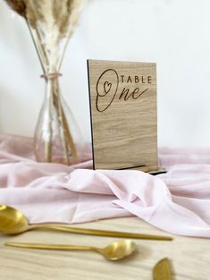 a wooden table number sitting on top of a pink cloth next to gold utensils