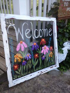 a welcome sign with flowers painted on the front and side of it, sitting next to a garden