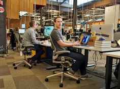 two men sitting at desks working on their computers in an open office space with large windows