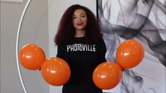 a woman holding orange balloons in front of her face and wearing a black shirt with white letters on it