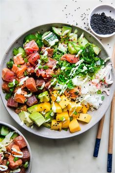 two bowls filled with food next to chopsticks on a white counter top and one bowl has salmon, avocado, and other vegetables in it