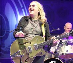 a woman singing into a microphone while holding a guitar in front of two other people