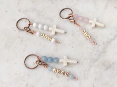 three different key chains with cross charms on them sitting on a marble surface, one is gold and the other two are white