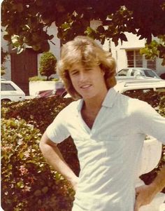 a woman standing in front of a bush with her hands on her hips, wearing a white shirt and shorts