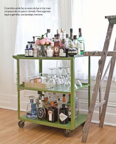 a green bar cart with bottles and glasses on it in front of a white window