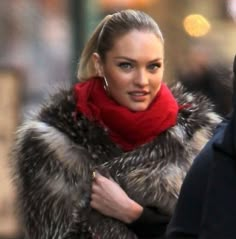 a woman in a fur coat and red scarf is walking down the street with her hand on her hip