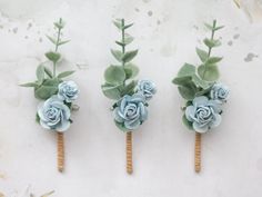 three boutonnieres with blue flowers and green leaves are arranged on a white surface