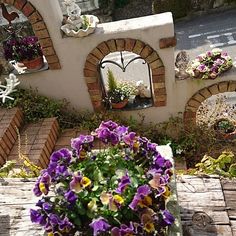 purple pansies are in front of a brick wall with an arched window and potted plants