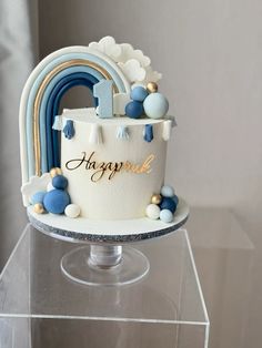 a birthday cake with blue and white frosting on a clear pedestal in front of a gray wall