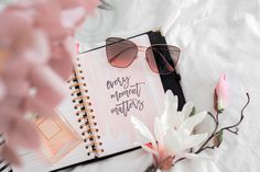 an open notebook with sunglasses on it next to pink flowers and a flower pot that says every moment matters