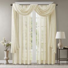 a living room with white curtains and a table in front of the window that has a lamp on it