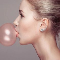 a woman blowing on a pink bubble with her nose close to the camera, against a gray background