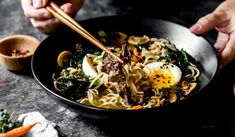 someone holding chopsticks over a bowl of noodles with meat, vegetables and eggs