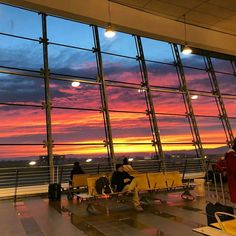 people are sitting on benches in front of large windows at an airport with the sun going down