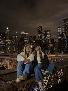 two young women sitting on a bridge in front of the city skyline at night time