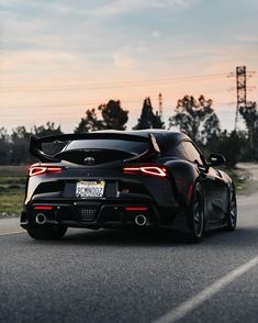 the rear end of a black sports car driving down a road at sunset with power lines in the background