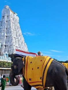 an elephant wearing a yellow blanket standing in front of a tall white structure with people walking around it