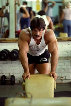 a man squatting down on a bench in a gym with other people working out behind him