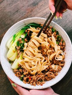 two hands holding chopsticks over a bowl of noodles and vegetables on a table